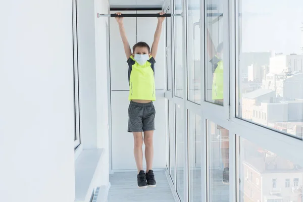 Boy in medical mask hang on horizontal bar on apartments balcony, doing sport exersise on quarantine, stay at home concept, self isolation during pandemic of coronavirus — Stock Image Boy Medical Mask Hang Horizontal Bar Apartments Balcony Doing Sport Stock Picture