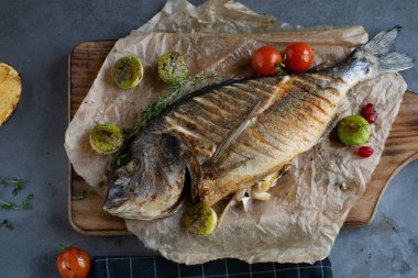 Yağlı kağıt üzerinde ızgara balık ve ahşap kesim tahtası. Etrafı ızgara domates ve kabakla çevrili.