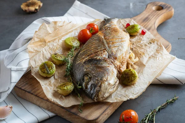 front view of grilled fish dorada on oiled paper and wooden cutting board   surrounded by grilled vegetables