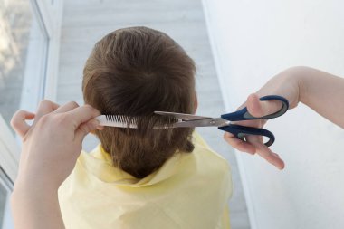 Kadın elleri bir tarak ve makas tutuyor ve çocuğun uzun saçlarını kesiyor, berber dükkanında berber dükkanı, aileler kuaförler kapalıyken saç kesiyor, evde kalıp konsept yapıyorlar. 