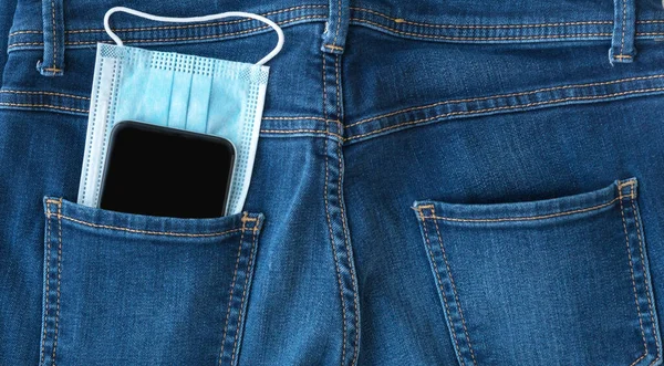 blue medical mask and black smartphone  look out from back  pocket of blue jeans, protection and prevention  measures from bacteria, viruses, coronavirus pandemic, need of social distance, healthcare concept