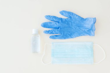 Koruyucu eşyaların üst görüntüsü, mavi tıbbi maske ve eldiven, arka planda beyaz bir şişe dezenfektan. Mesaj için yer var. Bakterilerden, virüslerden, koronavirüsten koruyucu ve önleyici önlemler. Sağlık konsepti, sosyal mesafeye ihtiyaç.