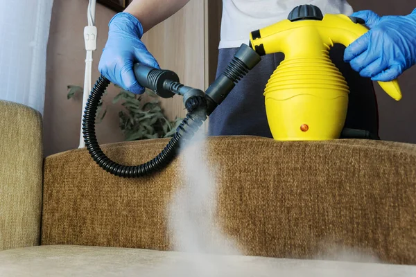 cleaning sofa with steam generator, hands in blue glove hold steam genetator and hose with brush, which produces steam for disinfection from microbes, dirt, allergy and viruses, home cleaning concept