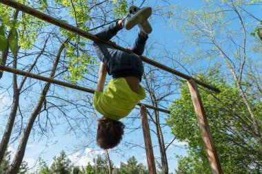  Çocuk büyük spor barlarında baş aşağı asılı duruyor. Ormanda büyük metal eski parmaklıklar var. Spor faaliyetleri her yerde, sağlık hizmetleri konsepti