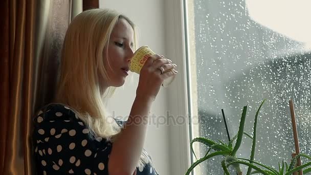 Closeup λυπημένος νέους κορίτσι πίνει καφέ στο σπίτι και κοιτάζοντας έξω από το παράθυρο — Αρχείο Βίντεο