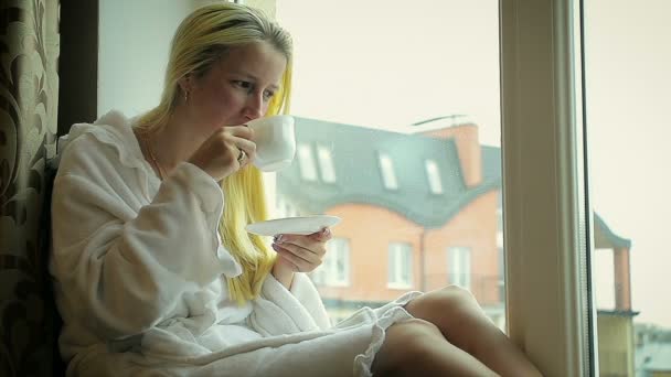 Junges Mädchen im Bademantel trinkt Kaffee zu Hause am Fenster — Stockvideo