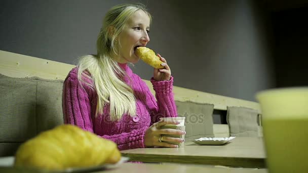 Junge Blondine isst Croissant und trinkt heißen Kaffee in einem Café — Stockvideo