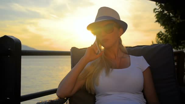 Chica joven feliz en un sombrero hablando en el teléfono inteligente en un viaje por el mar en la puesta del sol . — Vídeos de Stock