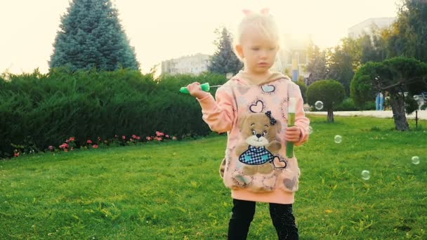 Movimiento lento: niña divertida de tres años soplando jabón-burbuja en el parque de la ciudad al atardecer . — Vídeo de stock