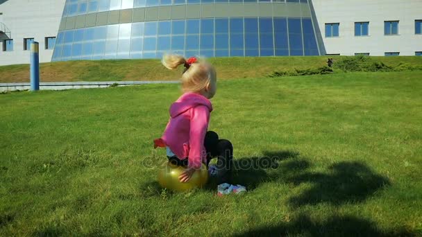 MOCIÓN LENTA: niña de tres años salta sobre una pelota de goma en un césped . — Vídeos de Stock