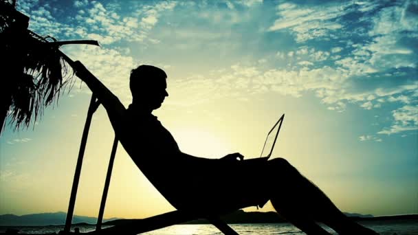 Siluett av mannen sitter på en stol och arbetar på en bärbar dator på en resort på solnedgången. — Stockvideo