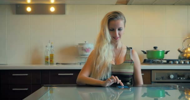 Dolly la giovane ragazza si siede a tavola e legge sullo smartphone in cucina a casa . — Video Stock