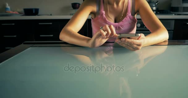 Chica joven se sienta a la mesa y leer en el teléfono inteligente en la cocina en casa . — Vídeos de Stock