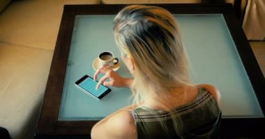 Dolly bir sarışın içecekler kahve ve mutfakta bir masada otururken telefonda bir makale okuma.