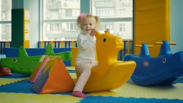 Movimiento lento: niña de tres años montando un caballo de juguete en un parque infantil en el interior . — Vídeos de Stock