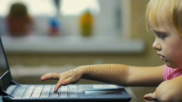 Dziewczyna z trzech lat powoli drukuje tekst na klawiaturze laptopa w domu w tle okna — Wideo stockowe
