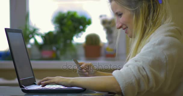 Glad ung blondin i en vit mantel håller ett kreditkort i hennes hand och gör inköp på Internet använder en bärbar dator. — Stockvideo