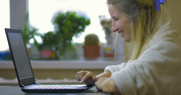 Fröhliche junge Blondine in weißer Robe wählt eine modische Tasche in einem Online-Shop zu Hause in der Küche. — Stockvideo