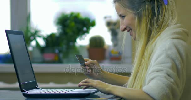 Närbild en glad blondin i en vit mantel håller ett kreditkort i hennes hand och gör inköp på Internet store använder en bärbar dator. — Stockvideo