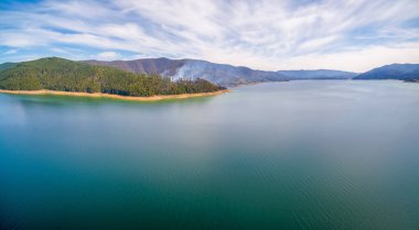 Ormandan yükselen duman bulutu göl kıyısındaki güzel manzaralı tepelerin arasından yükseliyor.