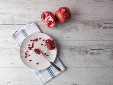 Nar bütün, yarısı ve tohumlar beyaz tabağa saçılmış, tahta kaşık ve kırsal masada çay havlusu. Kopyalama alanı olan üst görünüm.