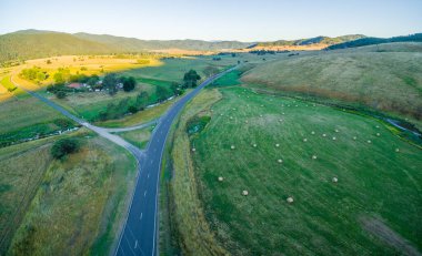Günbatımında Avustralya kırsalının hava manzarası. Yolun yakınındaki yeşil bir alana saman balyaları saçılmış..