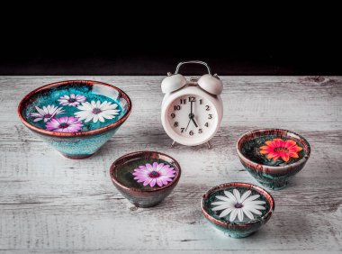 Su dolu ellerde yüzen Gerbera çiçekleri seramik kaseler ve kopyalanmış beyaz kırsal ahşap masa üzerinde antika bir çalar saat..