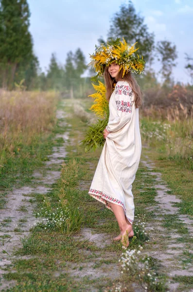 Jeune belle femme dans une couronne de fleurs sauvages et une ancienne tenue nationale marchant pieds nus sur le chemin . — Photo