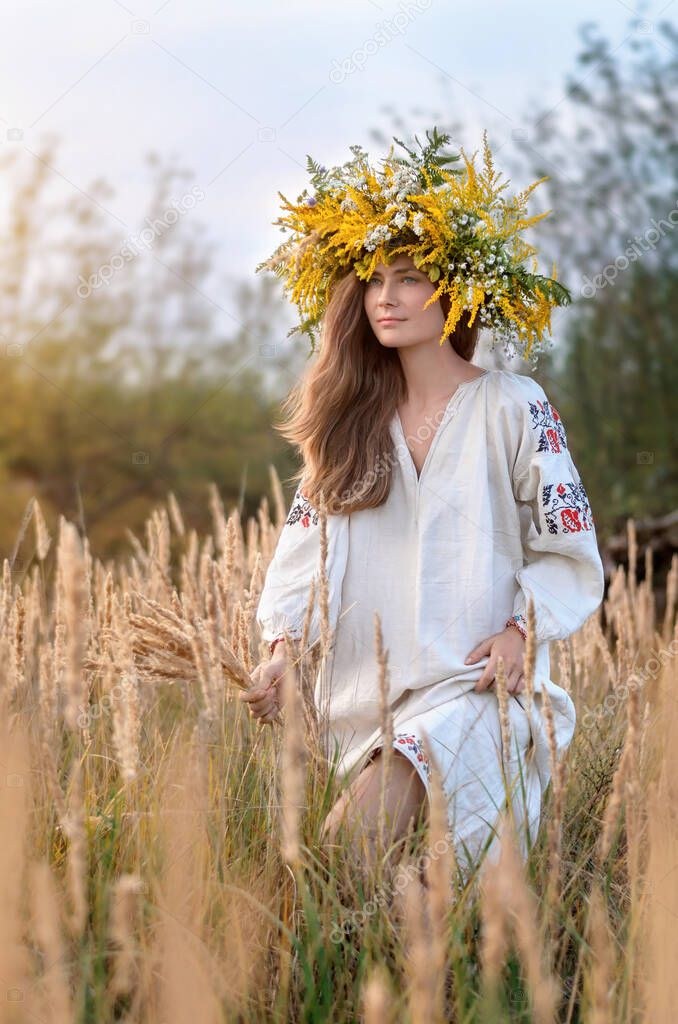 Young beautiful woman in a wreath of wildflowers and ancient embroidered shirt in a meadow of yellow dry grass.
