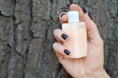 Siyah tırnaklı bir kadın eli ağaç kabuğunun arka planında bir şişe doğal kozmetik tutuyor.