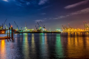 Hamburg 'daki kargo limanı gece aydınlandı. Konteynır terminalleri, kargo gemileri, vinçler ve açık mavi gökyüzü..