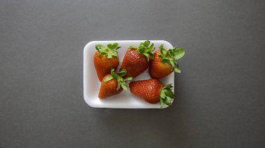 Gri bir arka plan ve sol altta yeşil yapraklı küçük kırmızı çilekler.