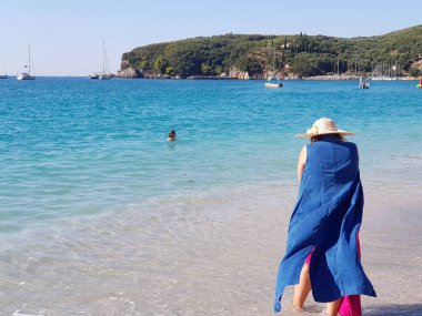 Hasır şapkalı, mavi elbiseli, kumsalda dinlenen ve Parga Yunanistan 'daki göle bakan genç kadının arka görüntüsü..