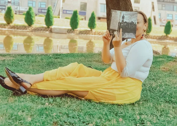 Skopje 北マケドニア 2019 若い素敵な気配りの女性は黄色のドレスで緑の草の上に横たわると都市公園で本を読む — ストック写真
