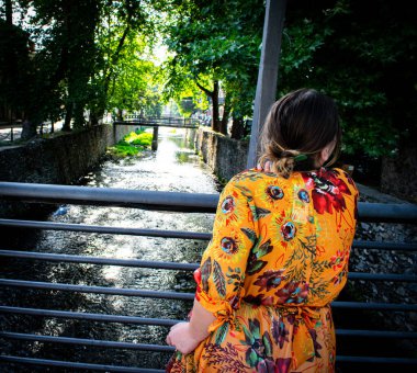 Artı, şehir meydanında tarihi unsurlar ve heykeller olan turuncu şık bir elbise giymiş ve poz vermiş bir model.