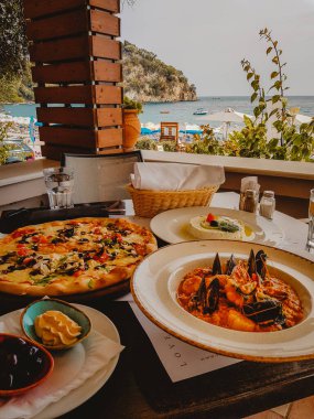 Tahta masada midyeli taze pizza ve makarna. Arka planda deniz manzaralı. 