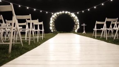 Düğün töreni için güzel bir kemer, geceleri ampullerin ışığı, beyaz sandalyeler ve beyaz ahşap bir yürüyüş yolu..