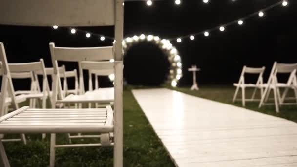結婚式のための美しいアーチ 電球からの光と夜 白い椅子と白い木製の歩道 — ストック動画