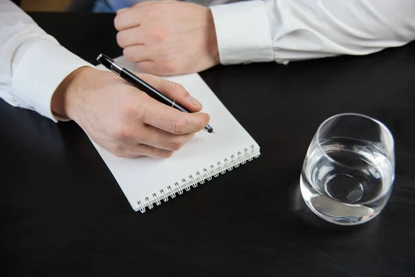 A man in a white shirt makes an entry in a notebook. Hands next to a notebook.