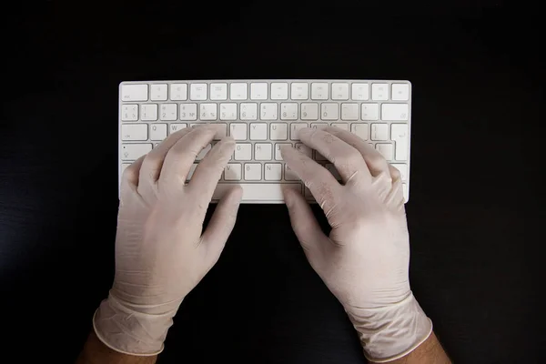Work during quarantine. Hands in rubber gloves on a white keyboard and black table. Work during quarantine.