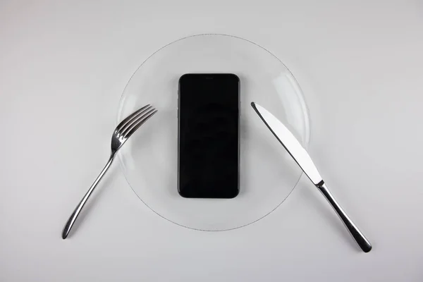 Şeffaf bir tabakta telefon olan demir çatal ve bıçak. Telefondaki bilgi tüketimi kültürü. Netiquette.