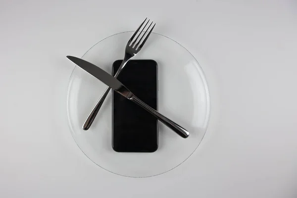 Şeffaf bir tabakta telefon olan demir çatal ve bıçak. Telefondaki bilgi tüketimi kültürü. Netiquette.