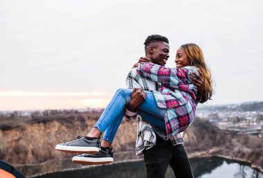 Koca, genç karısını güzel bir gökyüzünün arkasında kollarında tutuyor. Kareli gömlekler giyen şık bir çift..