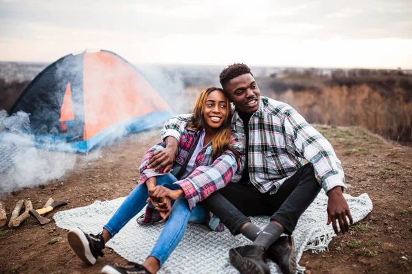 African American man and woman sitting on rug near campfire and tent, smiling, hugging each other. Man and woman in shirt.