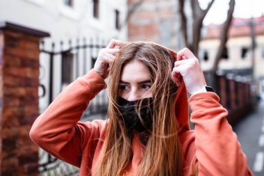 Siyah tıbbi maskeli kızıl saçlı genç bir kız koronavirüs salgını sırasında yürüyor. Salgın Coronavirus COVID-19. Yüz maskesi takan genç bir kız. Kapüşonlu kız. Kızıl kız..