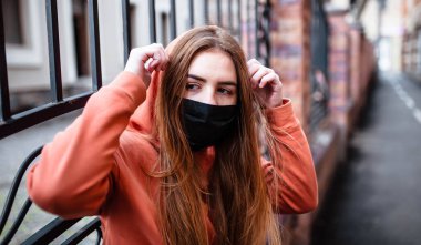 Siyah tıbbi maskeli kızıl saçlı genç bir kız koronavirüs salgını sırasında yürüyor. Salgın Coronavirus COVID-19. Yüz maskesi takan genç bir kız. Kapüşonlu kız. Kızıl kız..