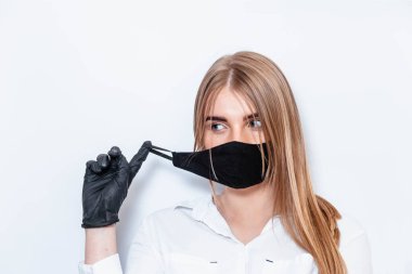 Beyaz takım elbiseli ve tıbbi maskeli bir doktor siyah eldivenlerle el ele tutuşup koronavirüs salgınının sonuçlarını düşünüyor. Beyaz arka planda bir doktor. Doktor siyah bir maske takıyor..