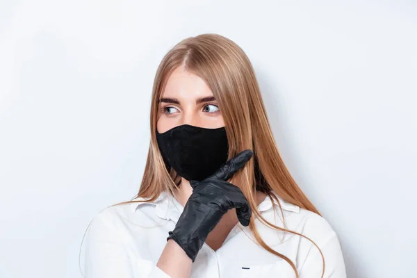 Beyaz takım elbiseli ve tıbbi maskeli bir doktor siyah eldivenlerle el ele tutuşup koronavirüs salgınının sonuçlarını düşünüyor. Beyaz arka planda bir doktor. Doktor siyah bir maske takıyor..