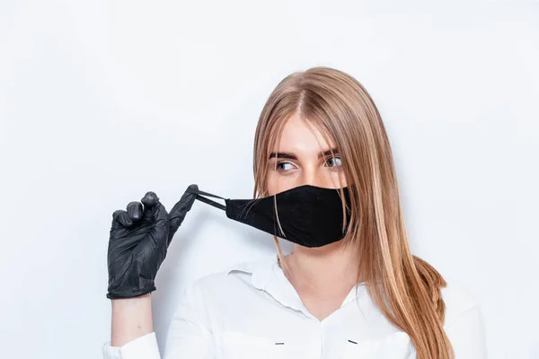 Beyaz takım elbiseli ve tıbbi maskeli bir doktor siyah eldivenlerle el ele tutuşup koronavirüs salgınının sonuçlarını düşünüyor. Beyaz arka planda bir doktor. Doktor siyah bir maske takıyor..
