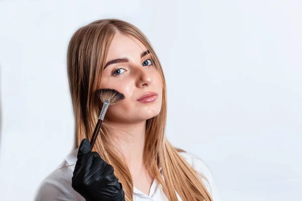 Beautiful young woman doing makeup. Girl with a brush in hand to apply powder on the face. Woman in black gloves makes makeup. Woman makes makeup herself on a white background.